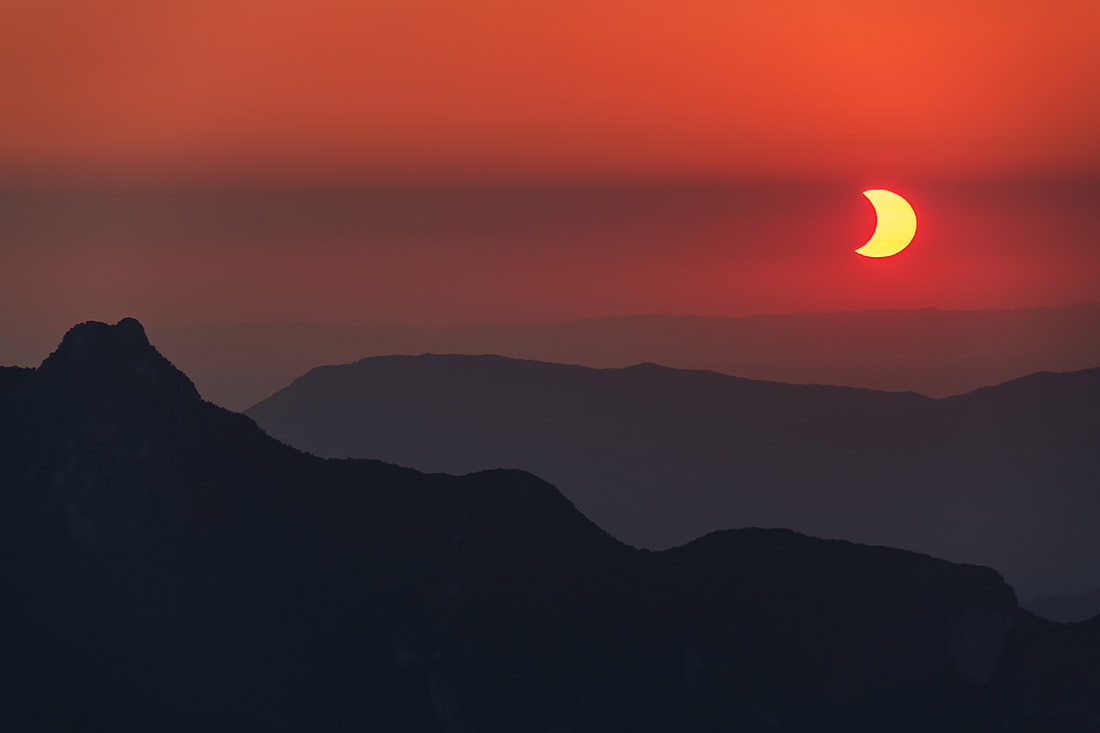 Croissant de soleil éclipsé dans un ciel rouge au-dessus de la silhouette de la Dent du Chat, le 12 août 2026